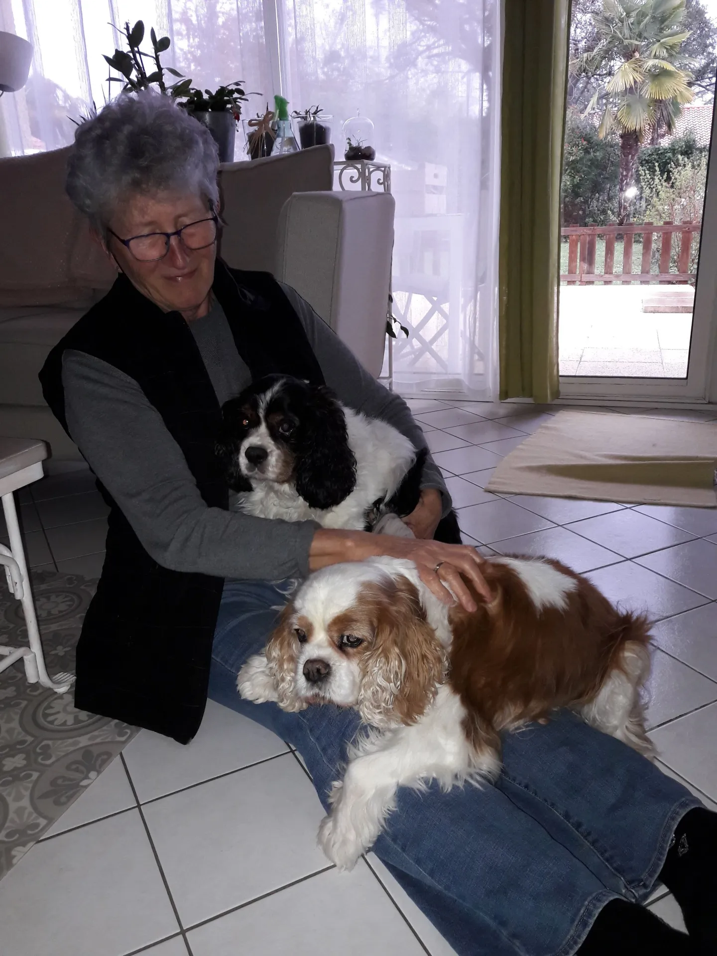 Annie assise au sol, deux cavaliers king charles sur les genoux, devant une baie vitrée ouverte sur un jardin