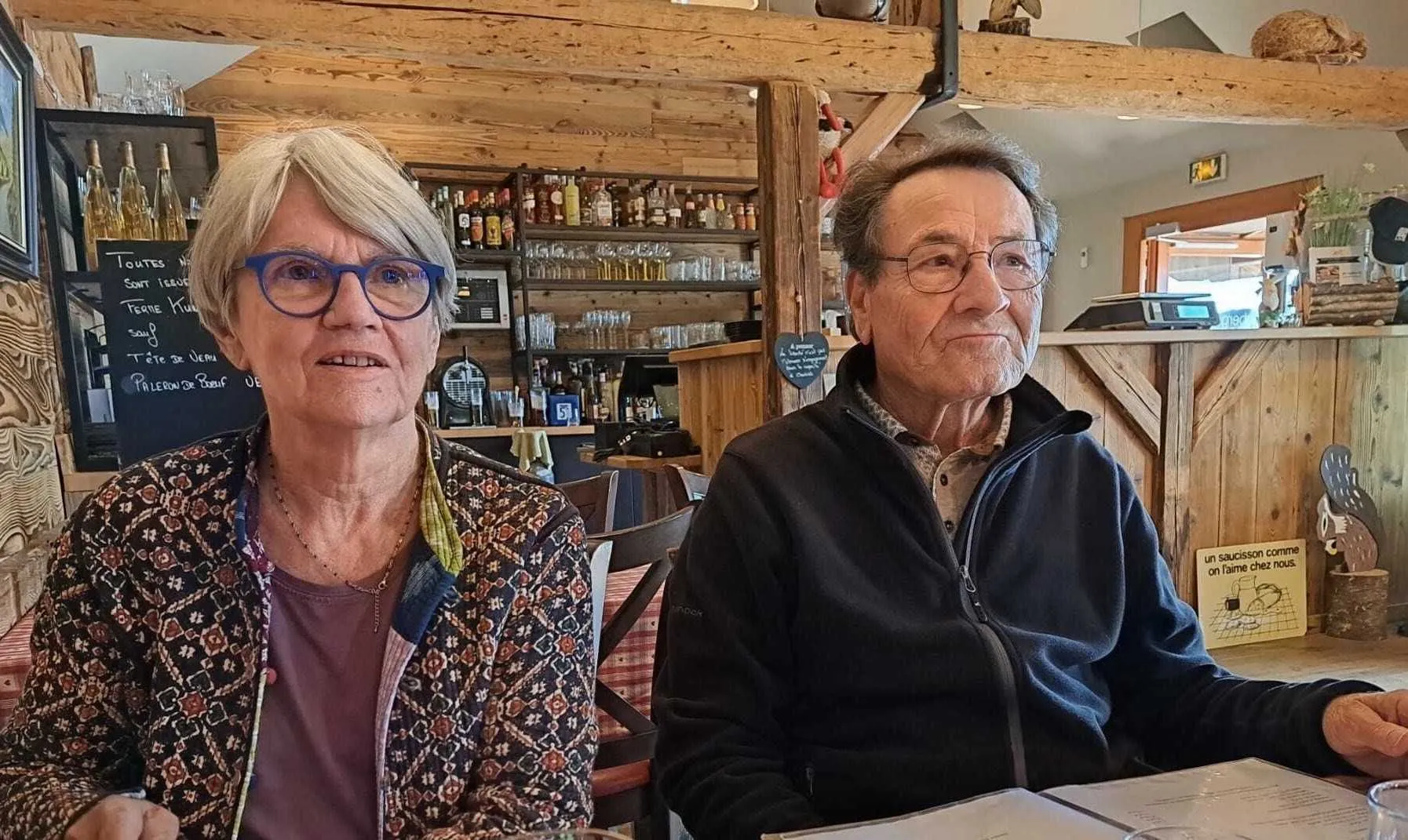 Josette et Michel attablés côte à côte dans un restaurant à la décoration en bois clair, le regard tourné vers la salle